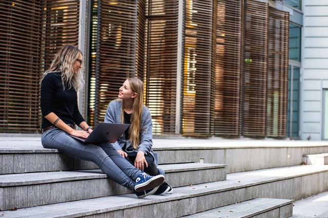 Zwei Frauen sitzen auf einer Treppe und unterhalten sich, eine hält einen Laptop